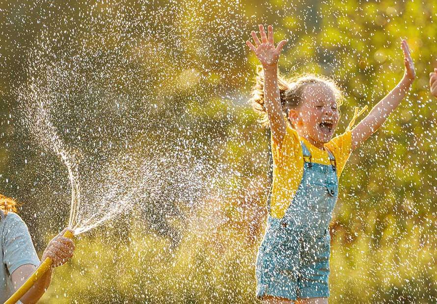mom spraying hose on jumping children in sun happy
